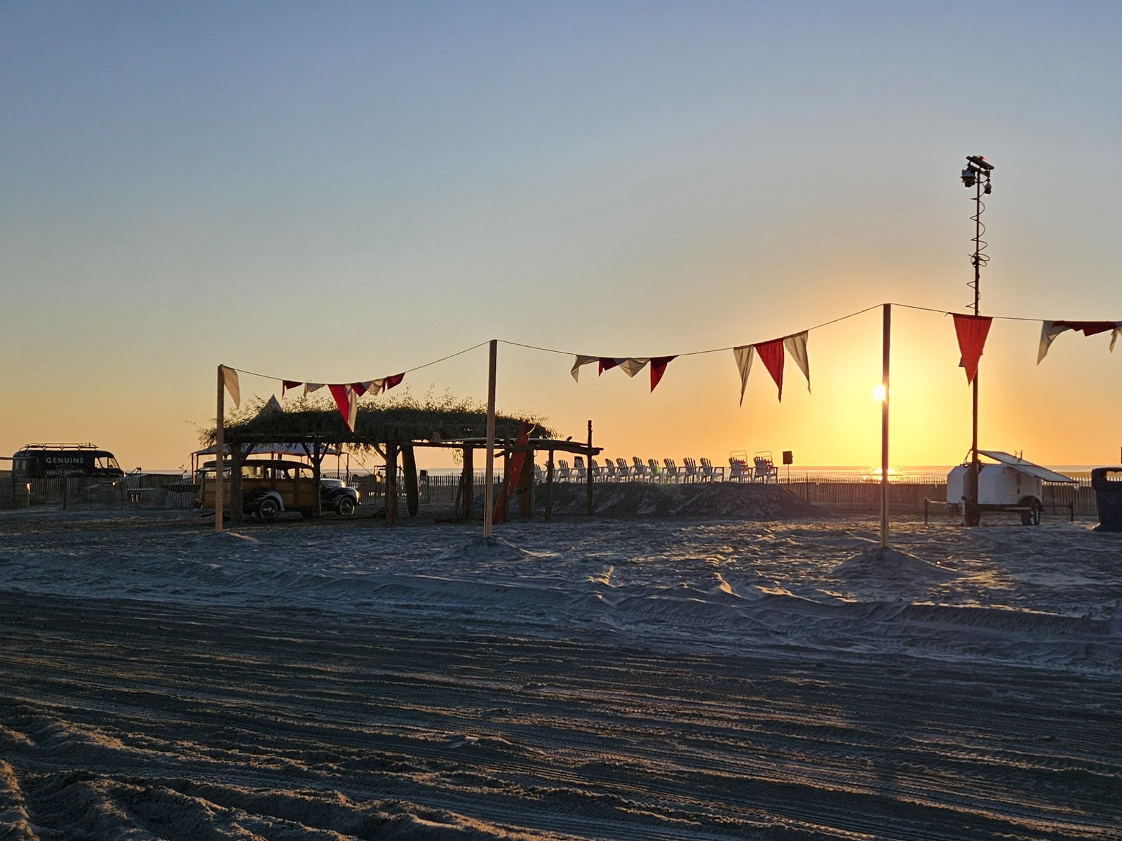 The beach drag strip at The Race of Gentlemen in Wildwood, New Jersey, graded flat at sunrise, with a wooden-bodied wagon under a thatched shelter and pennant bunting strung across the course.